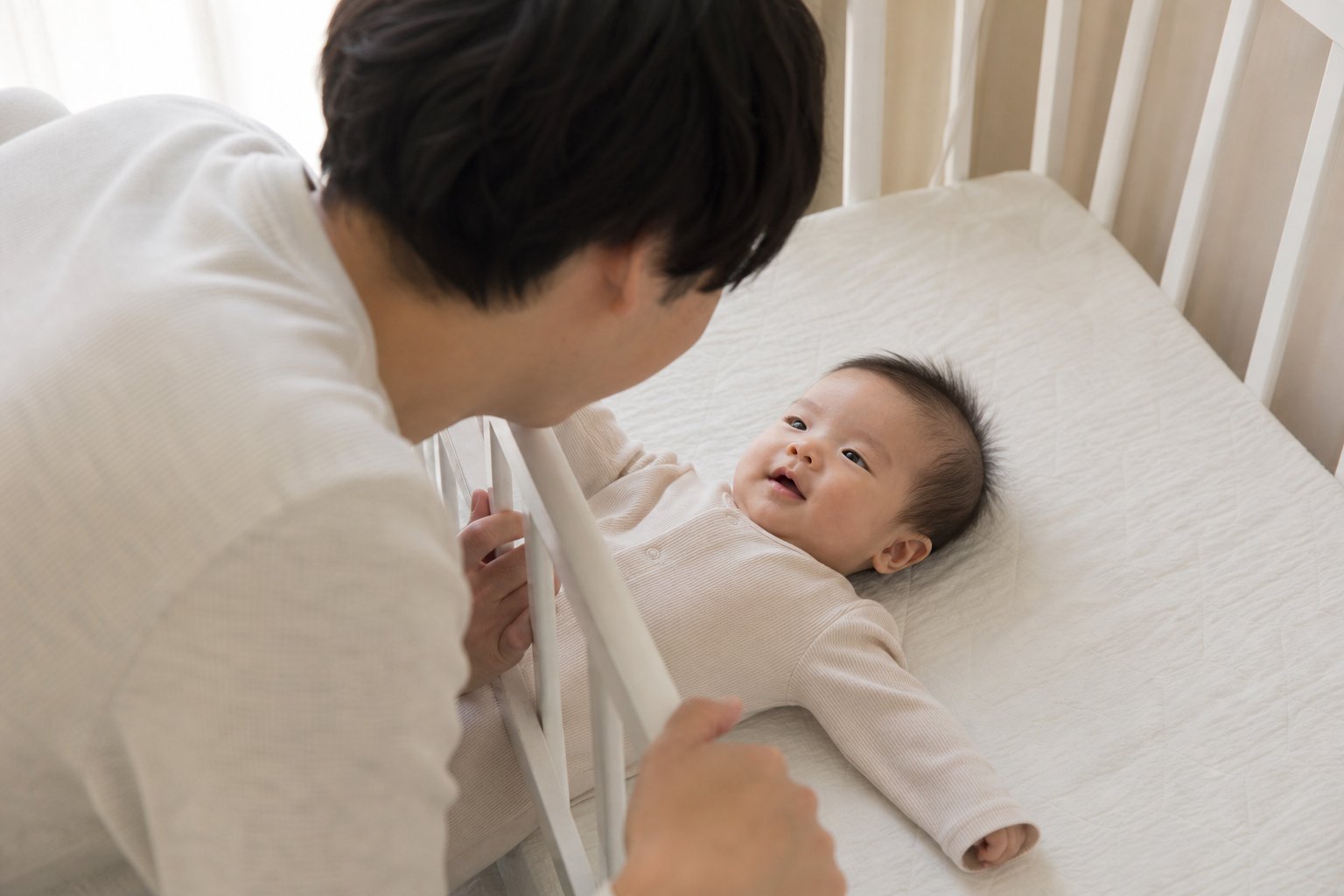 父親が白いベッド柵越しに、仰向けで笑う赤ちゃんの顔を覗き込む